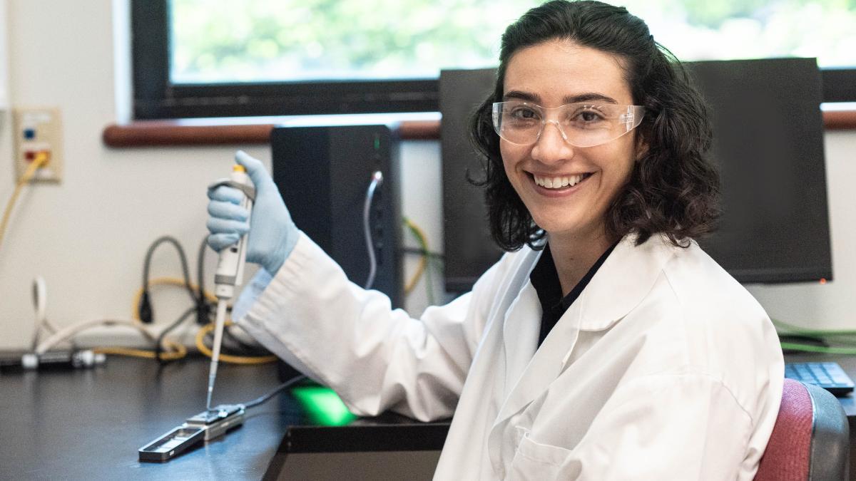 Erica Fuhrmeister smiles holding a micropipet in a lab.