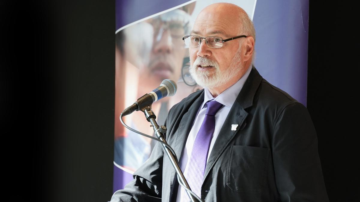 Mike Yost stands at a lectern, facing a crowd (not visible). He wears a dark jacket and purple tie. In the background is a tall banner.