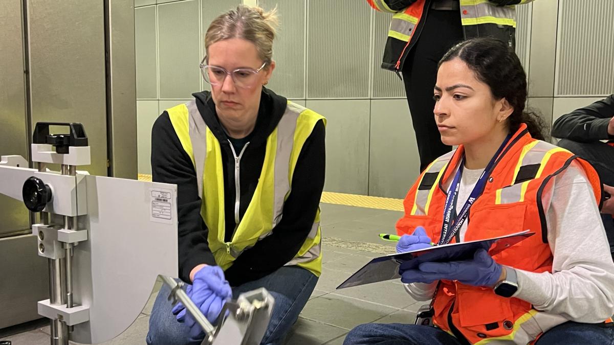 Two people in safety vests, one with blonde hair and glasses and the other with black hair, observe a measuring instrument at a transit station.