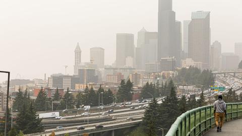 The Seattle skyline with hazy skies because of wildfire smoke. In the foreground of the image a person walks along an elevated walkway with a highway below.