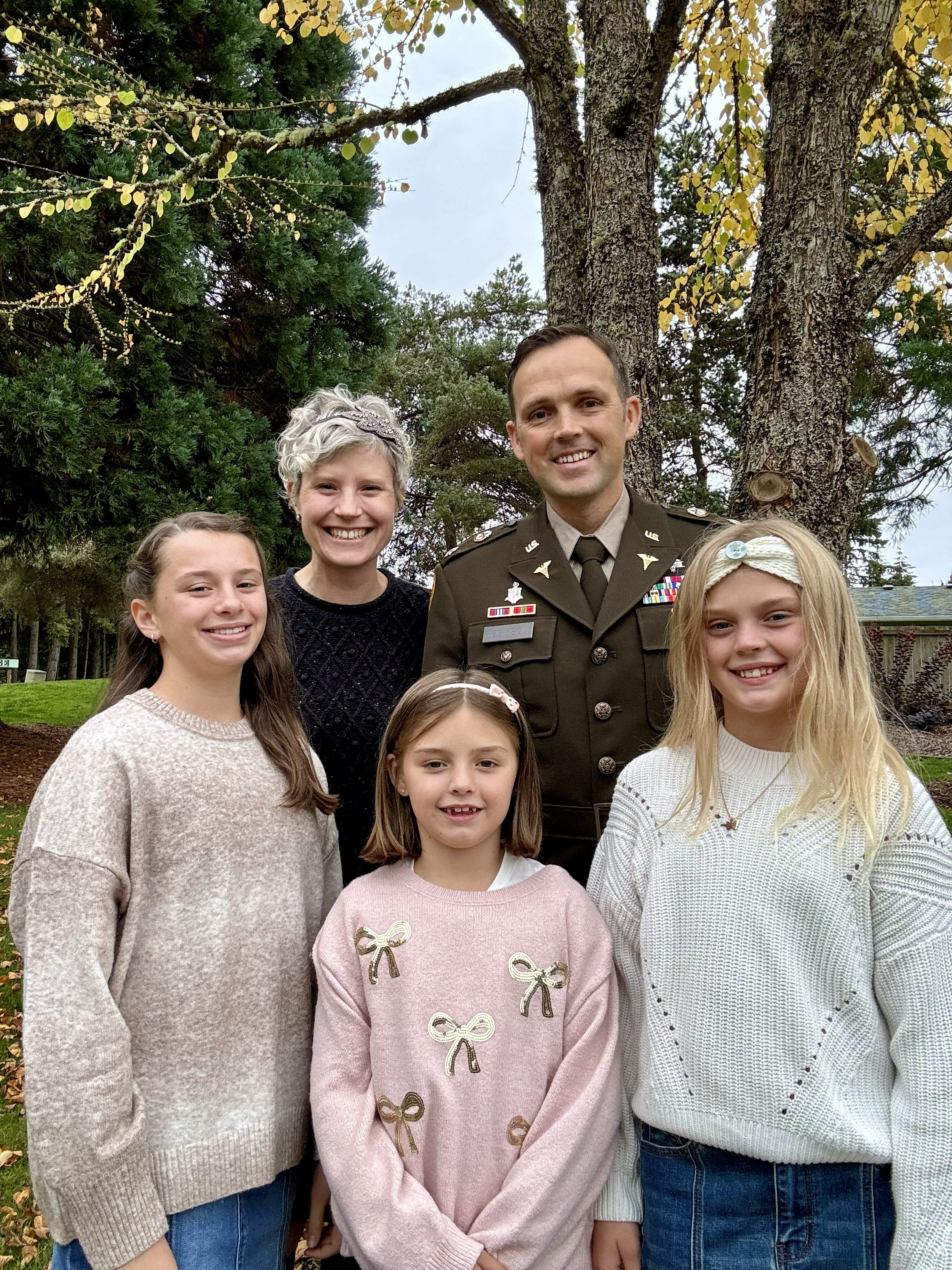 A family including a man in Army uniform (Mark Robinson), a woman and three children stand together smiling. Behind them is a deciduous tree with yellow leaves.