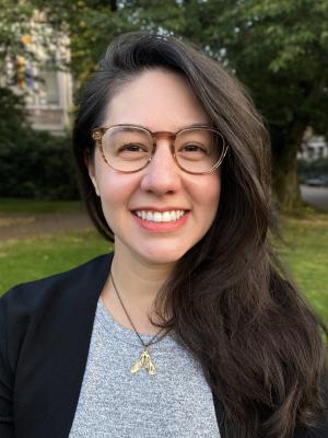 Headshot of Marie Spiker smiling and wearing glasses, standing outside with trees and a campus building behind her.
