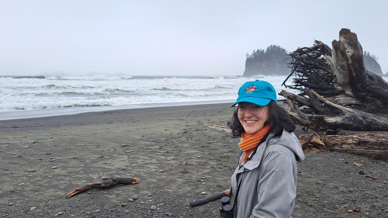 Martindale stands on a misty beach in front of large driftwood tree trunks, with sea stacks in the background. She is wearing glasses, a blue baseball cap, an orange scarf and a grey jacket.