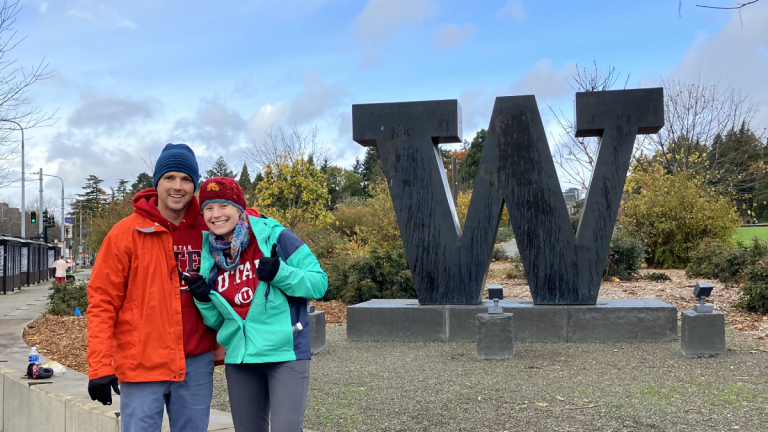 Mark Robinson and a woman stand together in front of a large W sculpture on the campus of the University of Washington. 