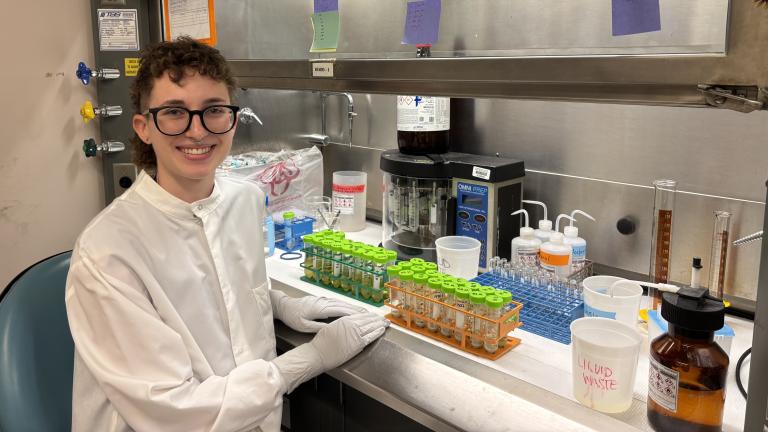 Person with short curly brown hair and glasses sits in a lab near a counter full of test tubes and other equipment.