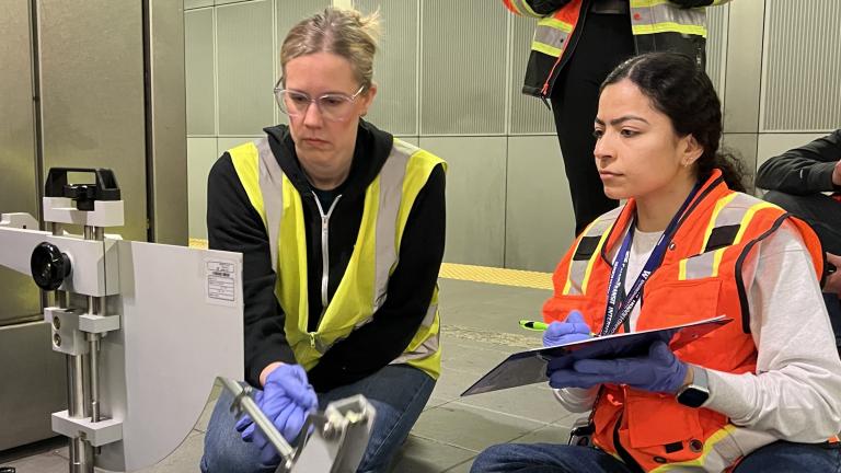 Two people in safety vests, one with blonde hair and glasses and the other with black hair, observe a measuring instrument at a transit station.