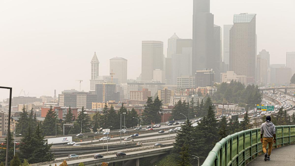 The Seattle skyline with hazy skies because of wildfire smoke. In the foreground of the image a person walks along an elevated walkway with a highway below.