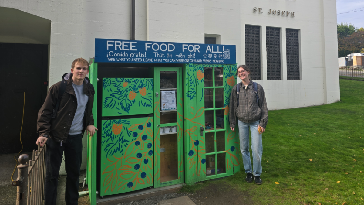 Two people stand on either side of an outdoor refrigerator and food storage cabinet. A sign above the community fridge and micropantry reads "Free food for all! (translated into Spanish, Vietnamese and Chinese) Take what you need leave what you can we're one community, friends + neighbors." The micropantry is painted green with flowers and is in front of a church labeled "St. Joseph Parish." 