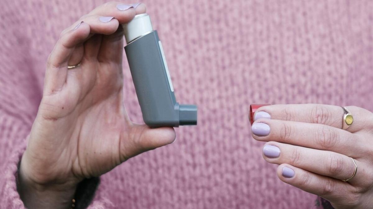 Person wearing pink sweater holds an asthma inhaler in one hand and red cap in the other.